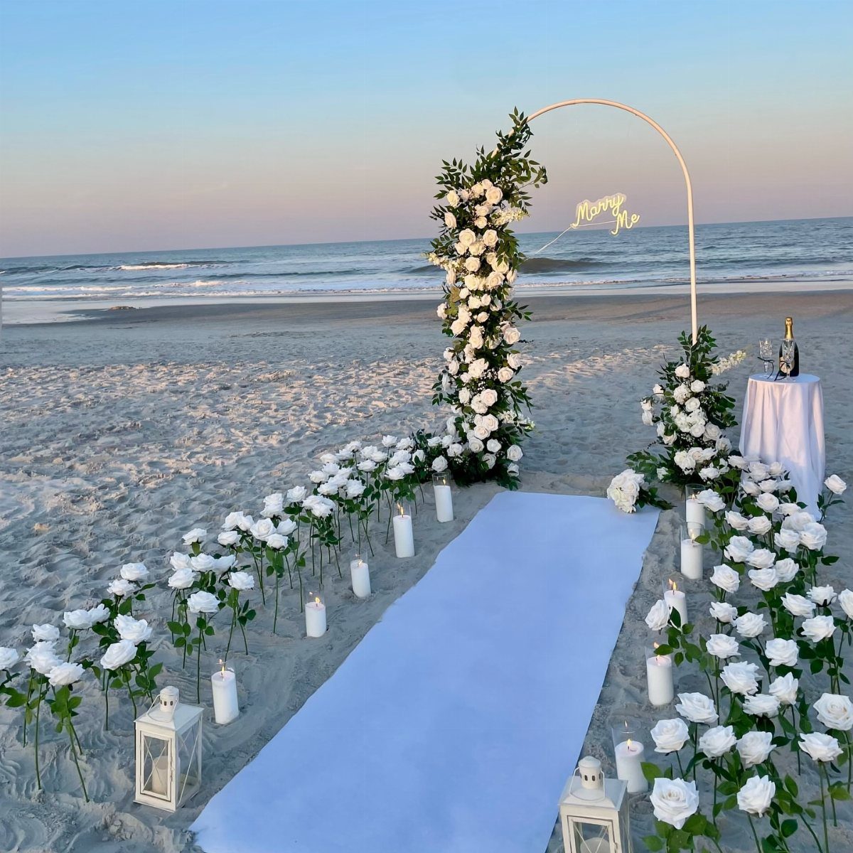 Beach proposal setup with white roses, candles, and a floral arch with neon sign.