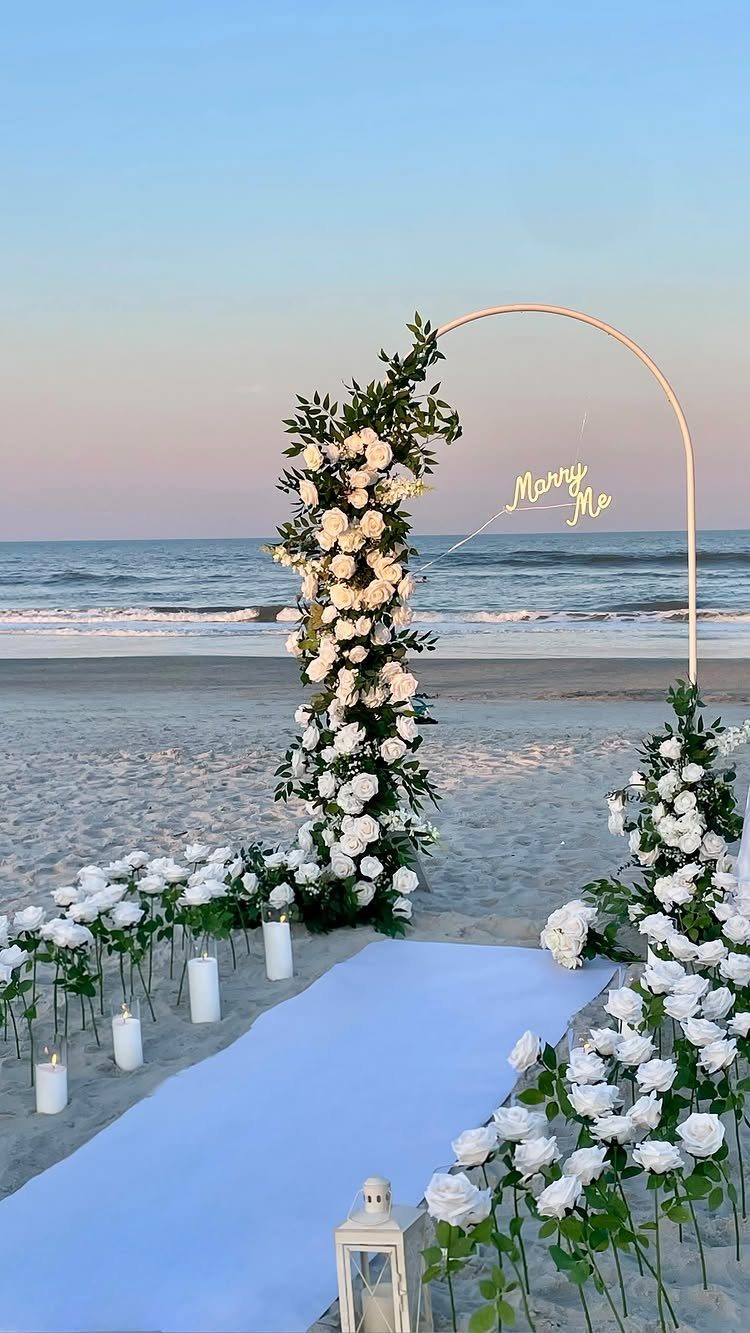 Gold circular arch with white roses and candles on a sandy beach at sunset.