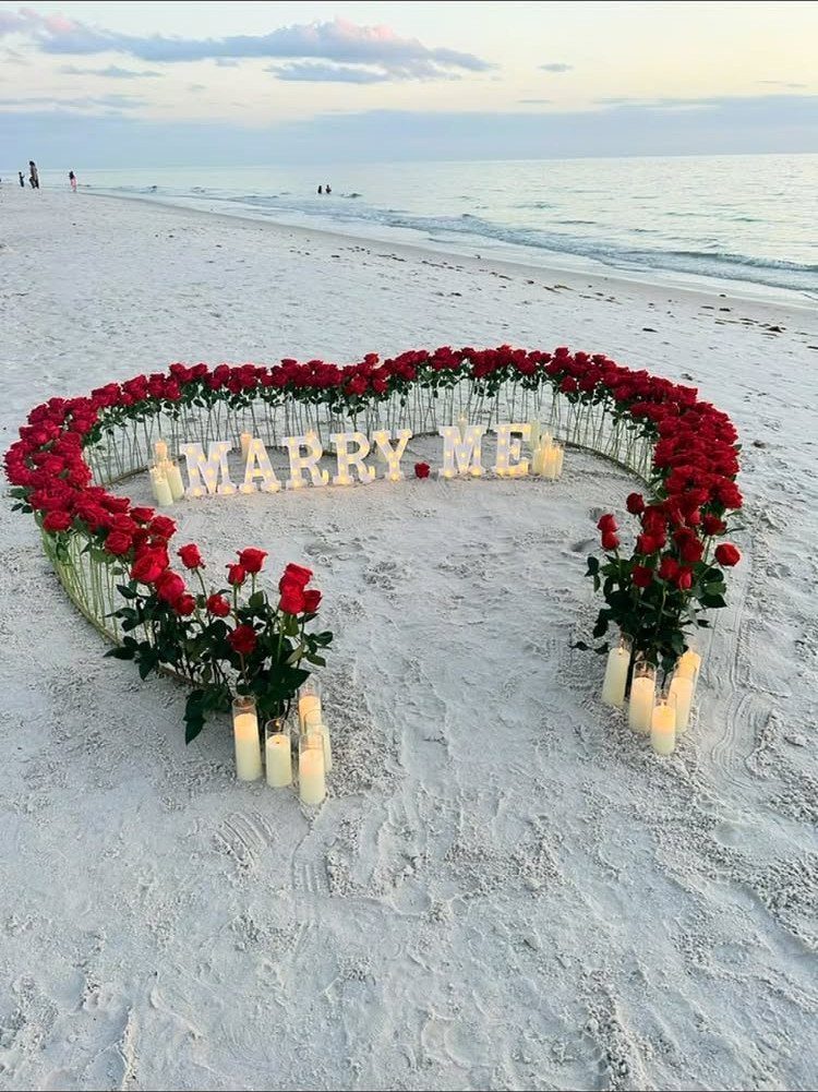 Heart-shaped rose petal arrangement with Marry Me letters on a white sand beach.