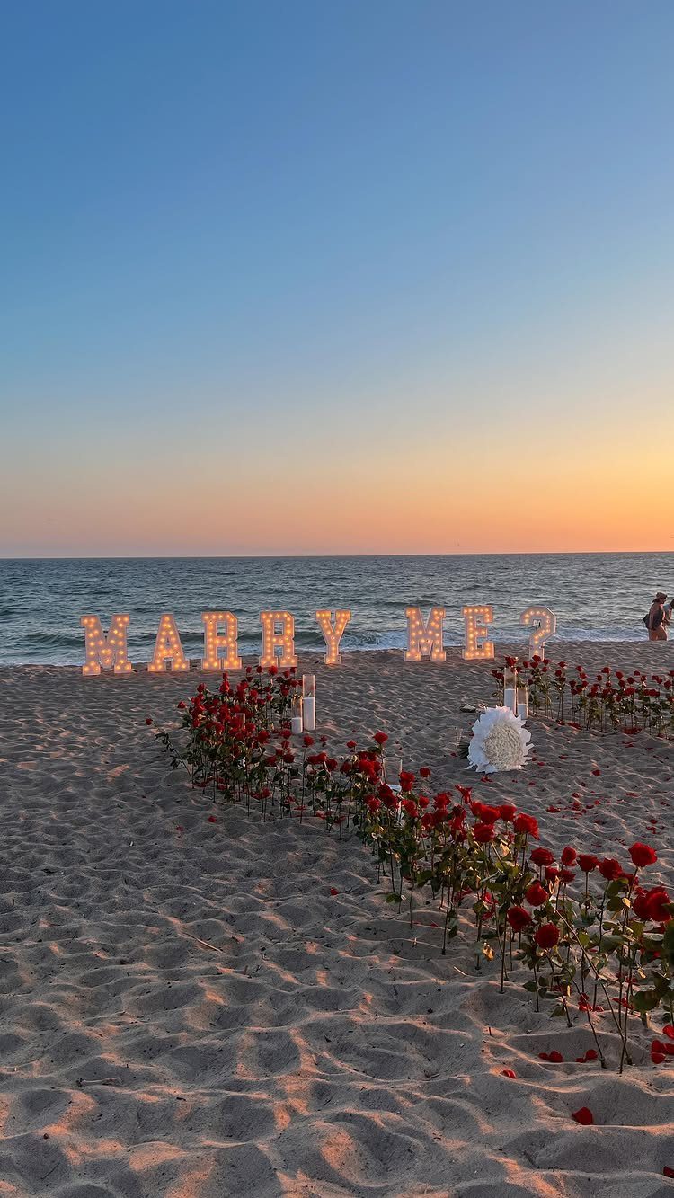 Illuminated Marry Me letters and red roses in a heart on a beach.