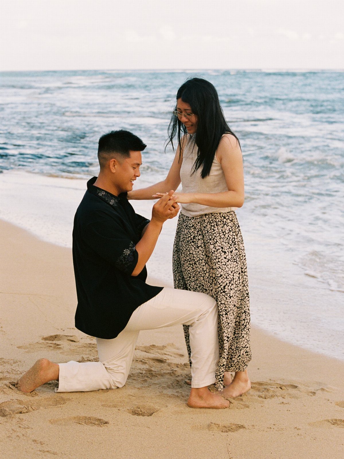 Man kneeling on a sandy beach proposing to a woman by the ocean.