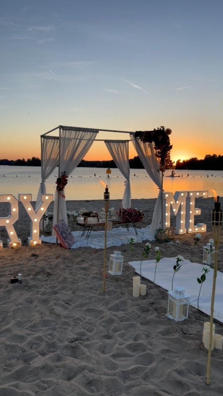 Romantic beach proposal setup with illuminated letters, white canopy, and picnic blankets at sunset.