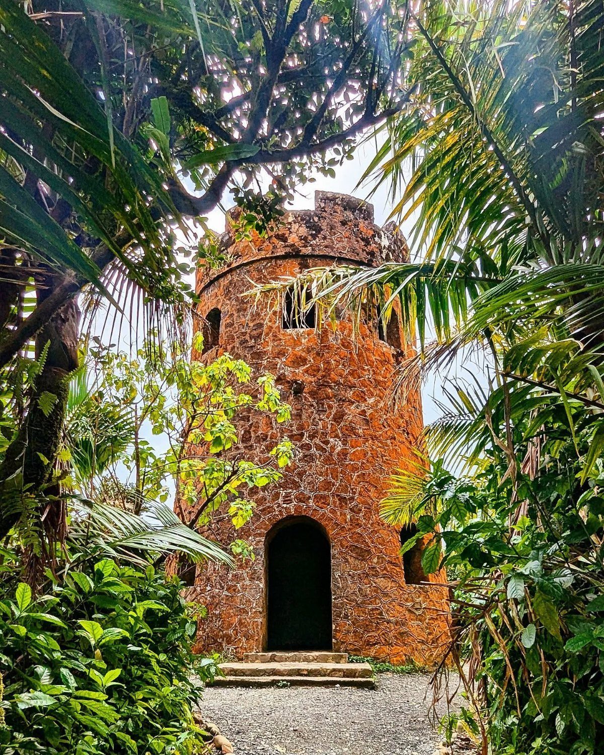 Reddish-brown stone tower surrounded by lush tropical greenery and palm trees.