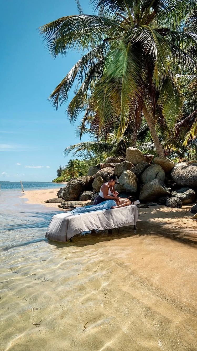 Couple relaxing on a beach lounge bed under palm trees by turquoise water.