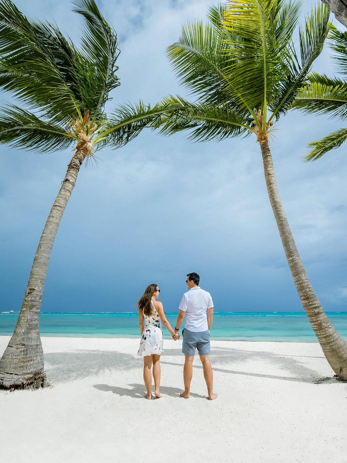 Couple holding hands on a white sand beach framed by two palm trees.