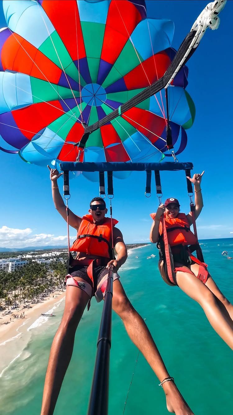 Couple parasailing over a tropical beach with a colorful rainbow parachute above them.