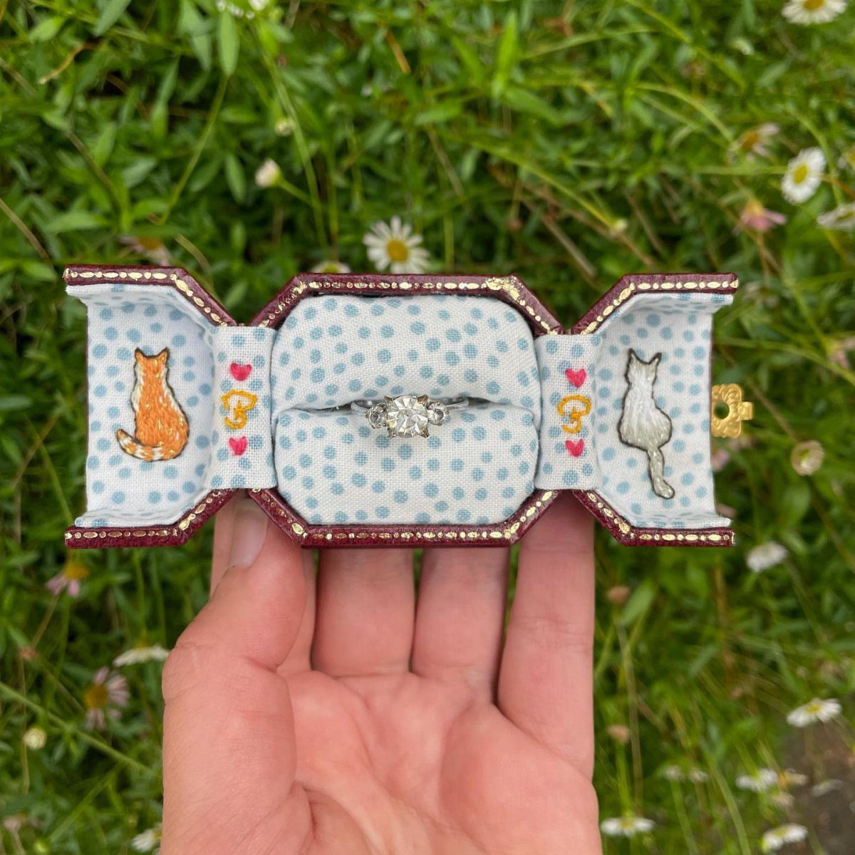 Diamond ring inside an embroidered cat-themed ring box held over green grass.