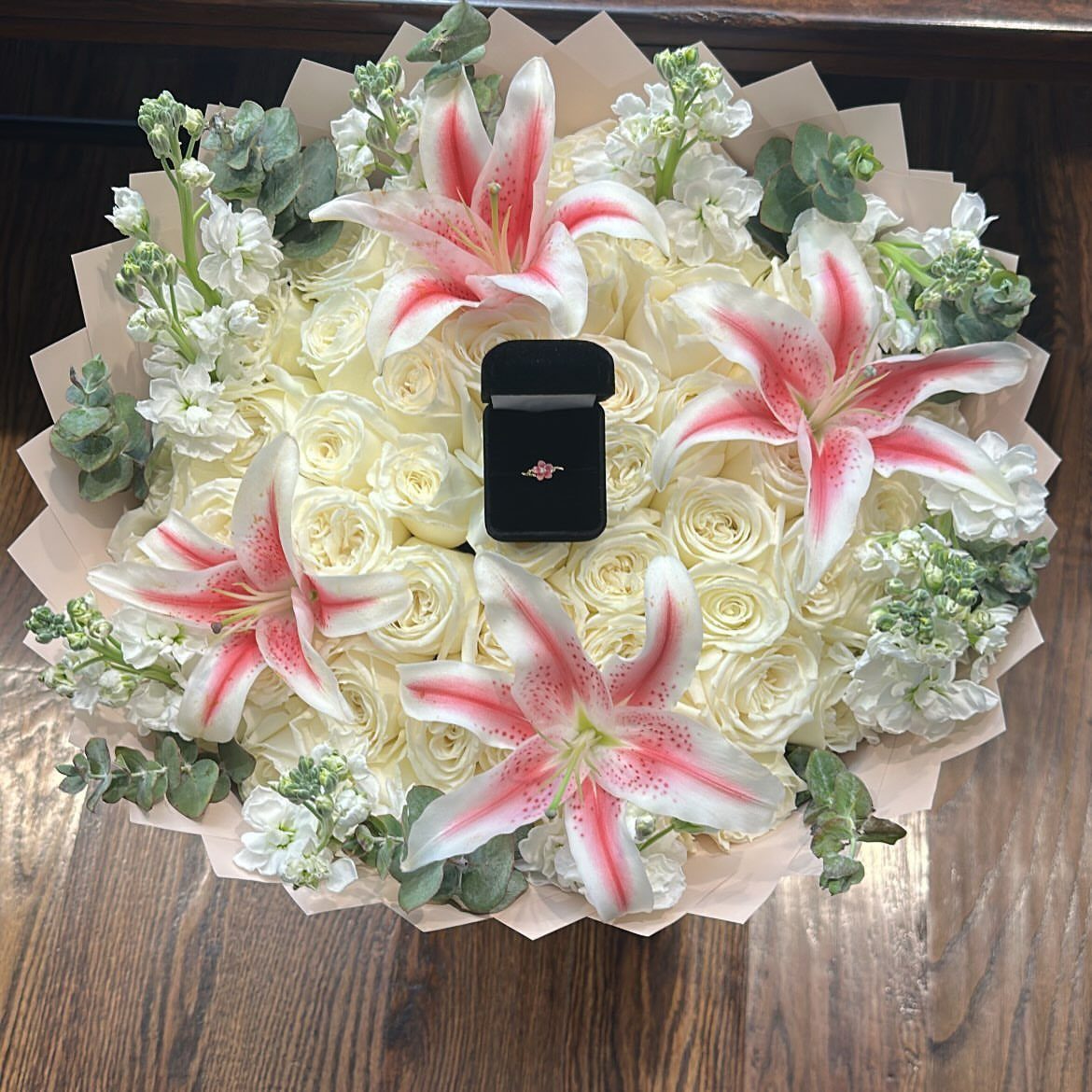 Gold ring in a black box resting on white roses and pink lilies.