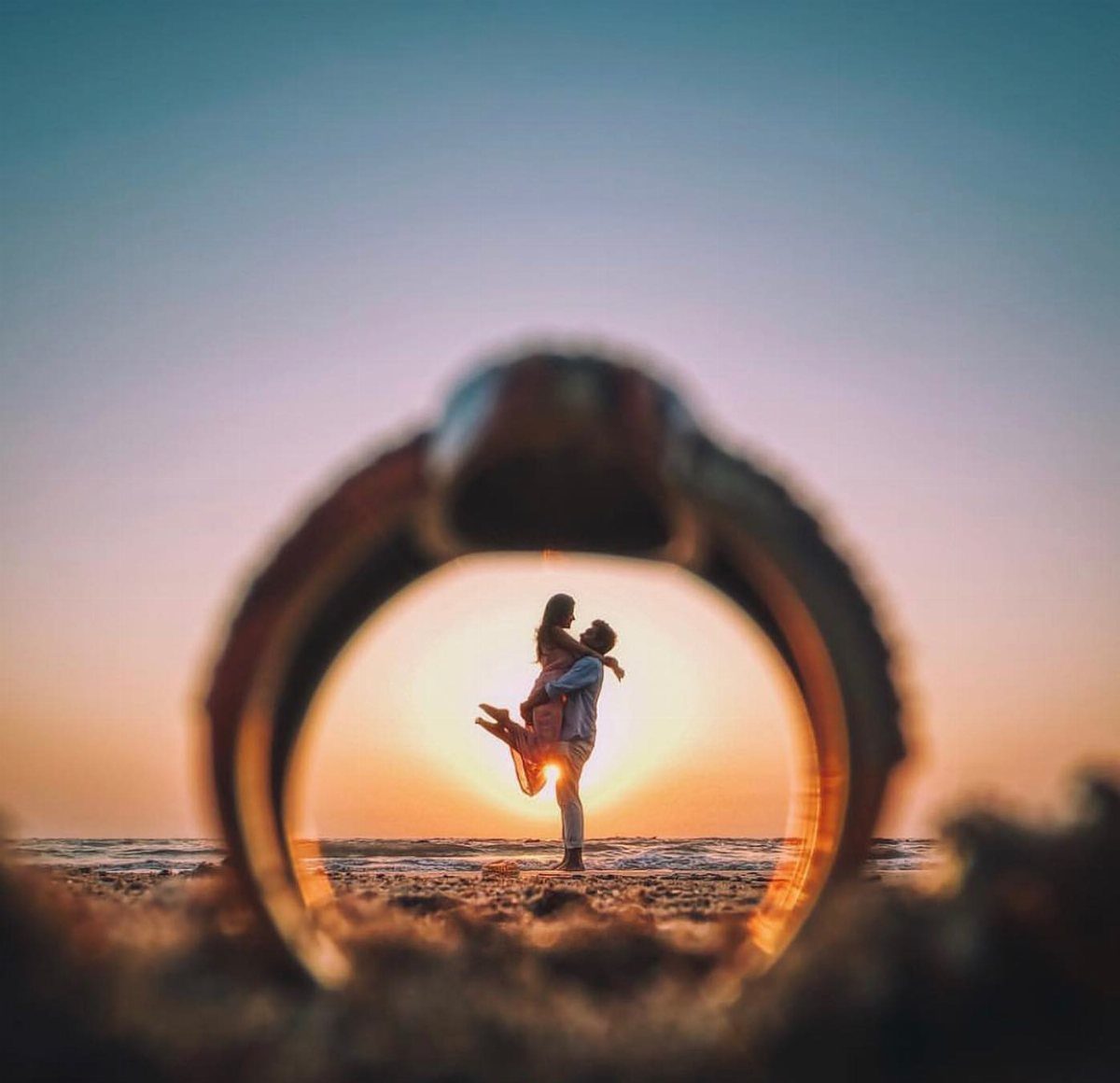 Wedding ring framing a silhouetted couple embracing on a beach at sunset
