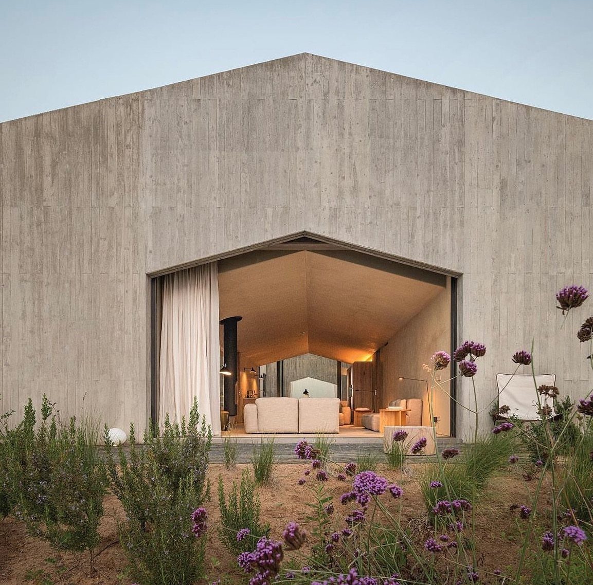 Modern concrete villa with open living area surrounded by purple wildflowers and sand.