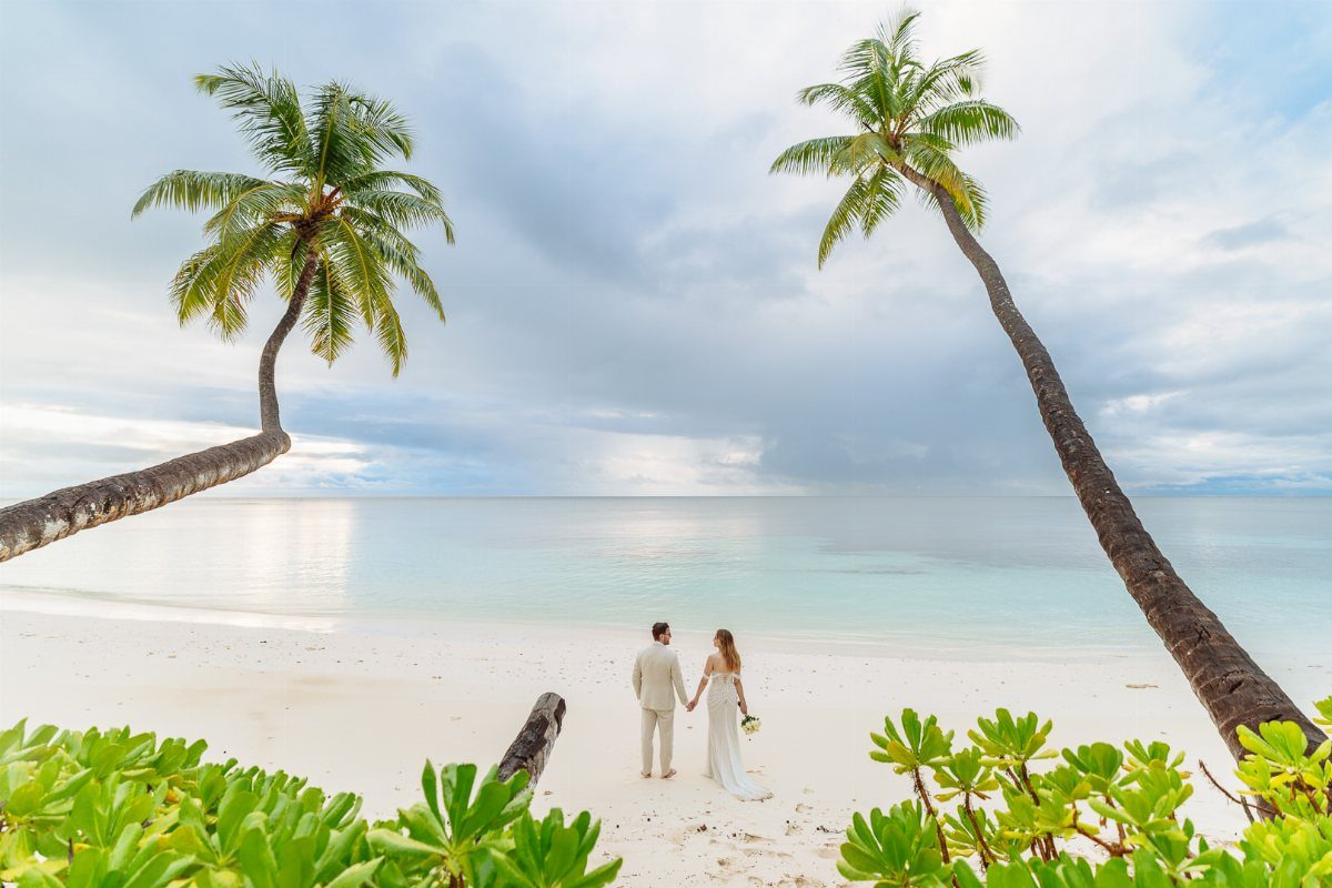 Newlywed couple holding hands on a tropical white sand beach with leaning palm trees.