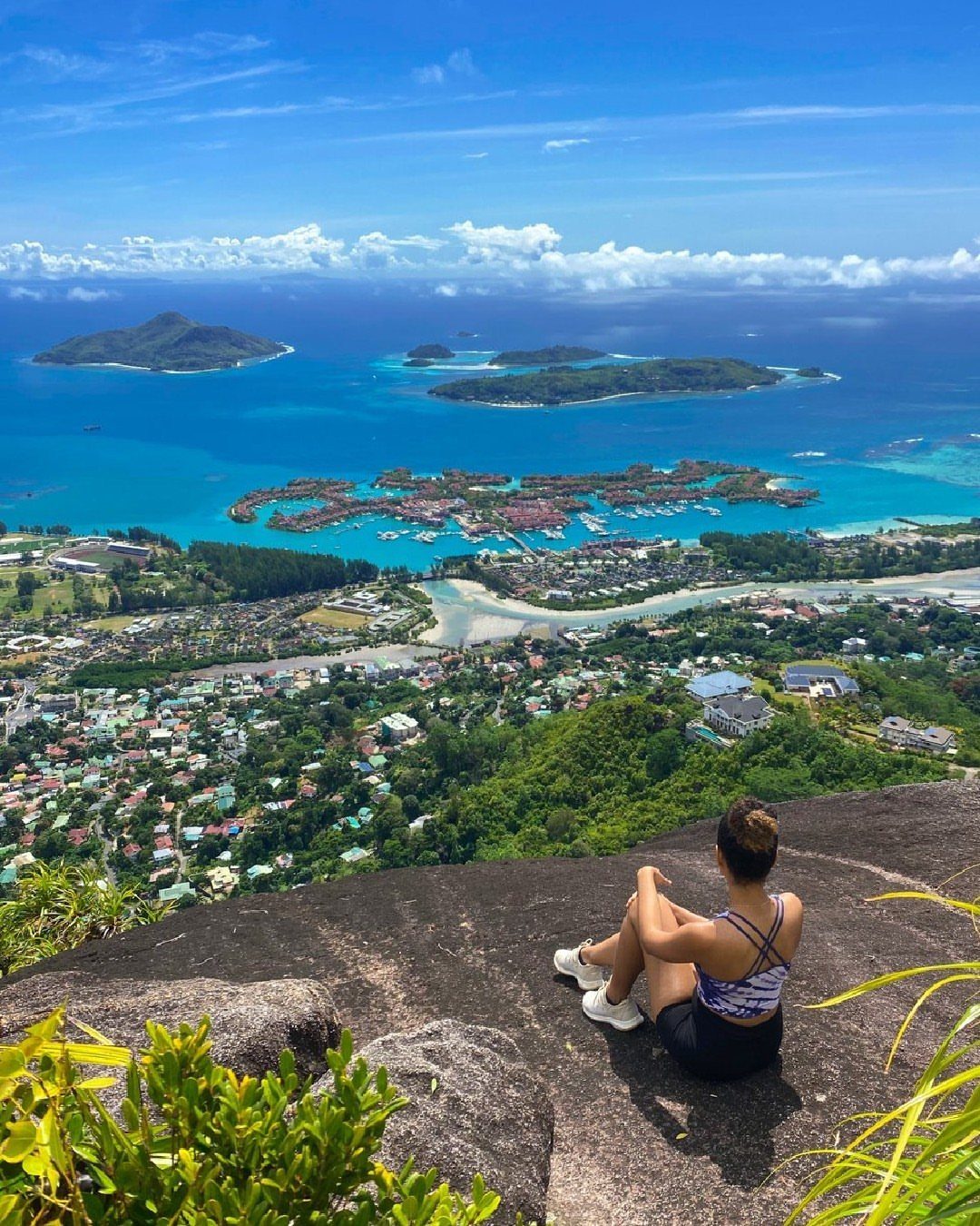 Woman sitting on a rocky cliff overlooking a tropical coastal town and islands.