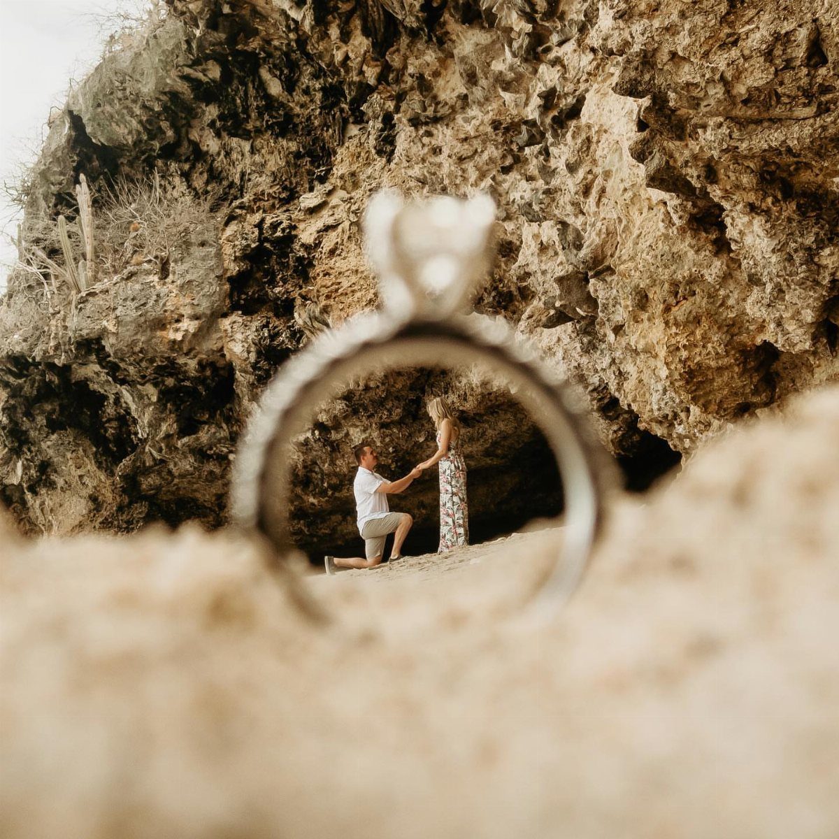 Engagement ring framing a man proposing to a woman on a sandy beach.