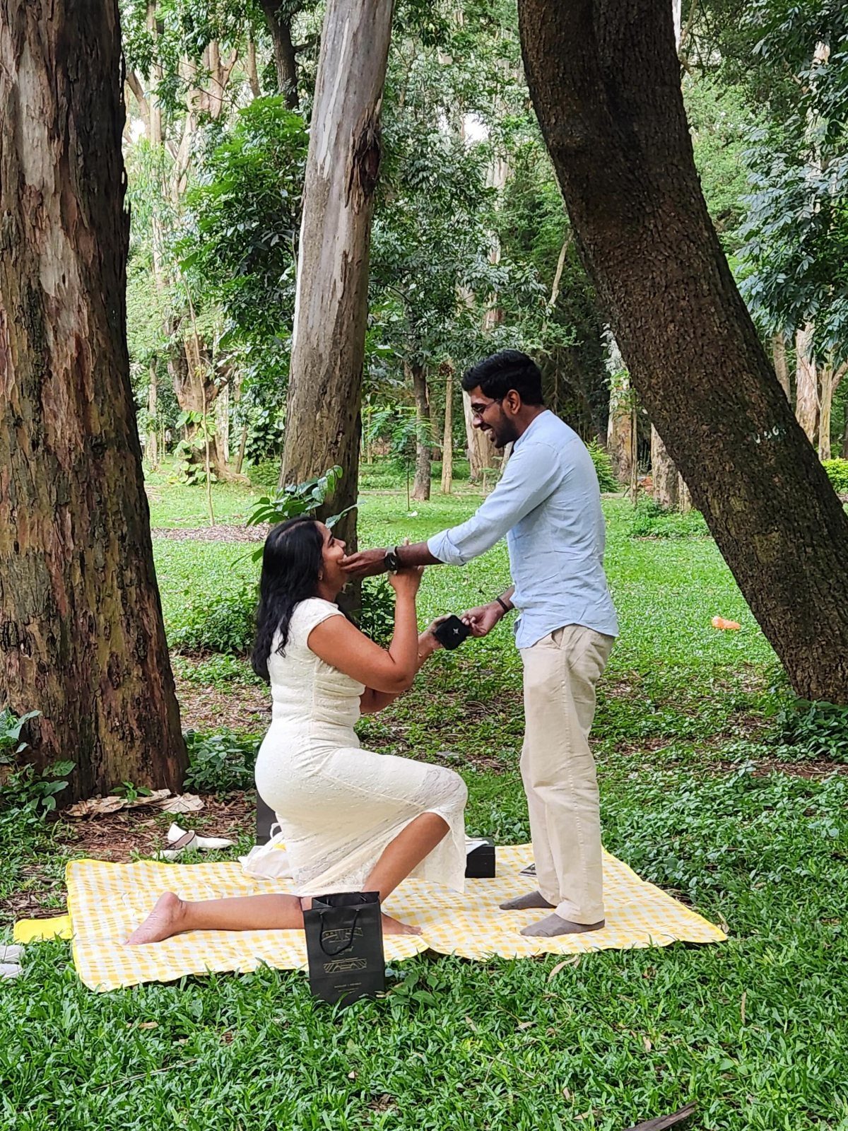 Woman kneeling on a mat proposing to a man in a wooded park.