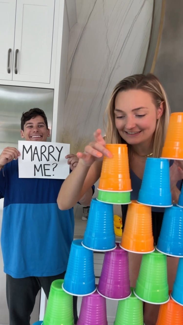 Woman stacking colorful plastic cups while a man holds a Marry Me sign behind her.