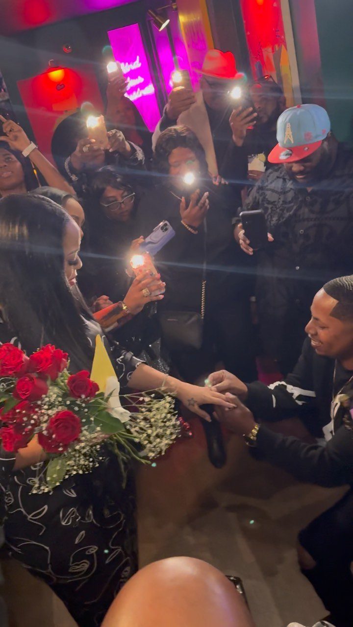 Man proposing to a woman with red roses surrounded by cheering friends filming.