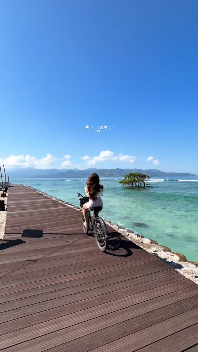 Woman in white dress riding a bicycle on a wooden boardwalk by turquoise water