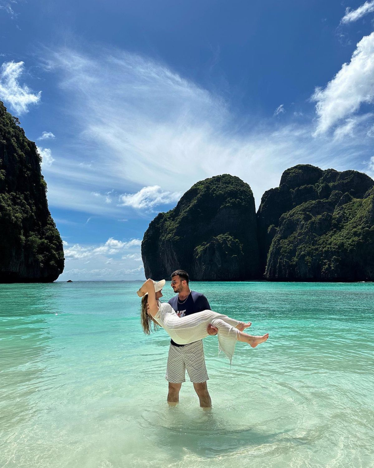 Couple embracing in turquoise water on a beach with towering limestone cliffs.