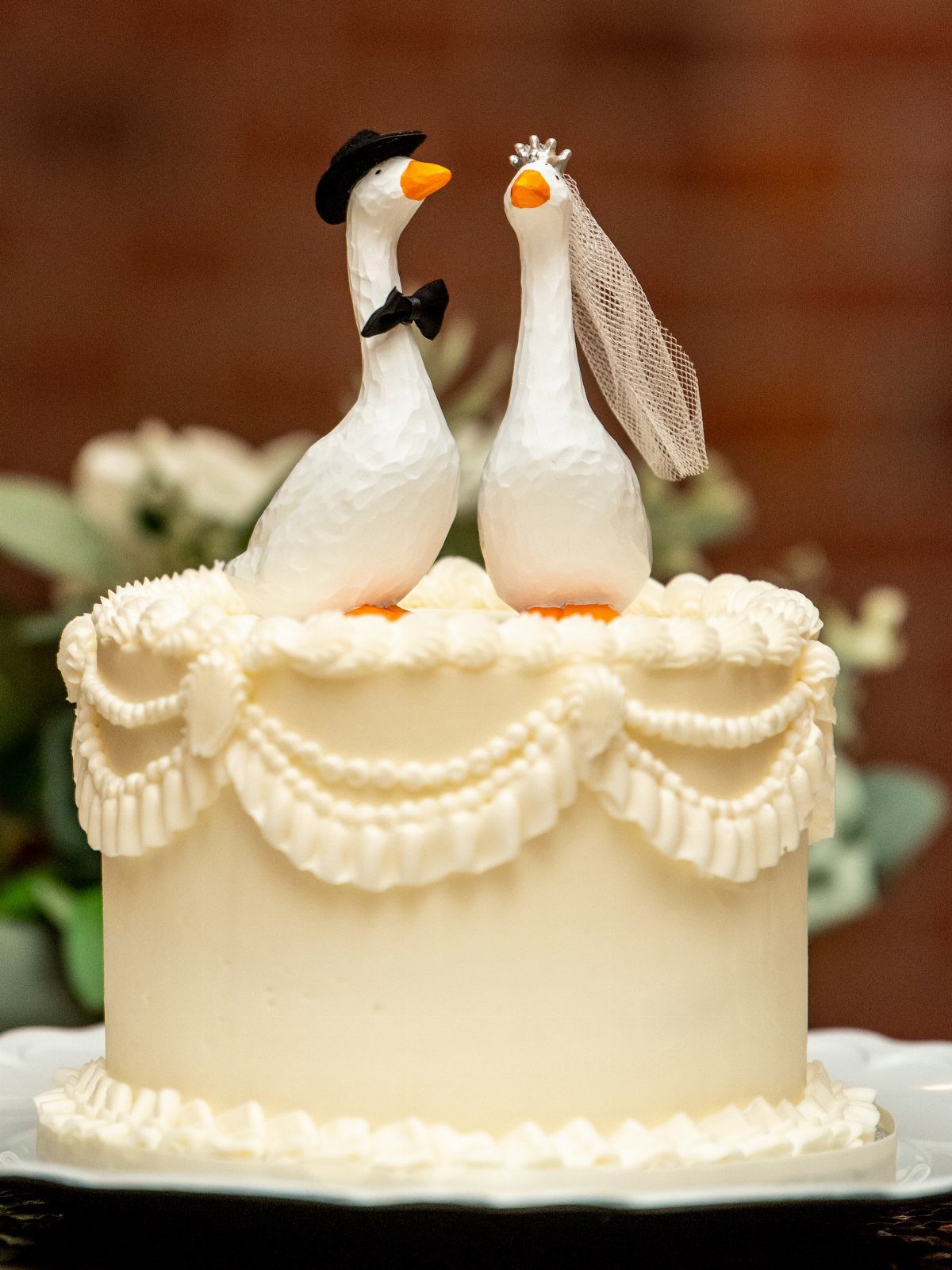 White wedding cake with ornate piping topped with two dressed goose figurines.