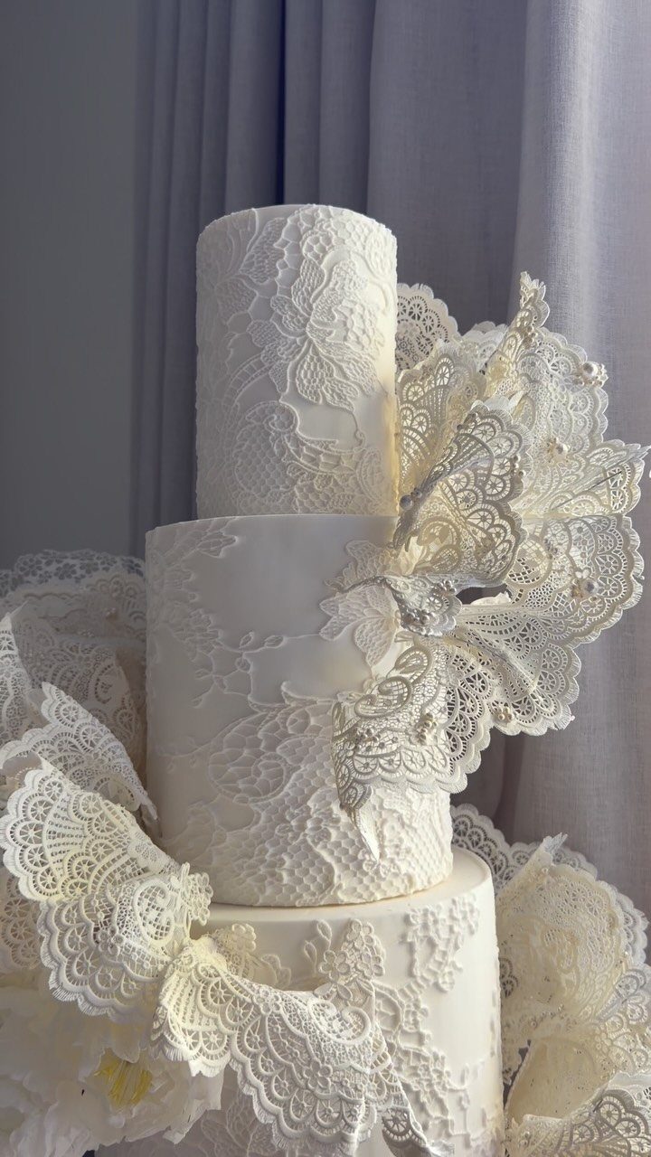 Three-tiered white wedding cake adorned with intricate edible lace and floral lace decorations.