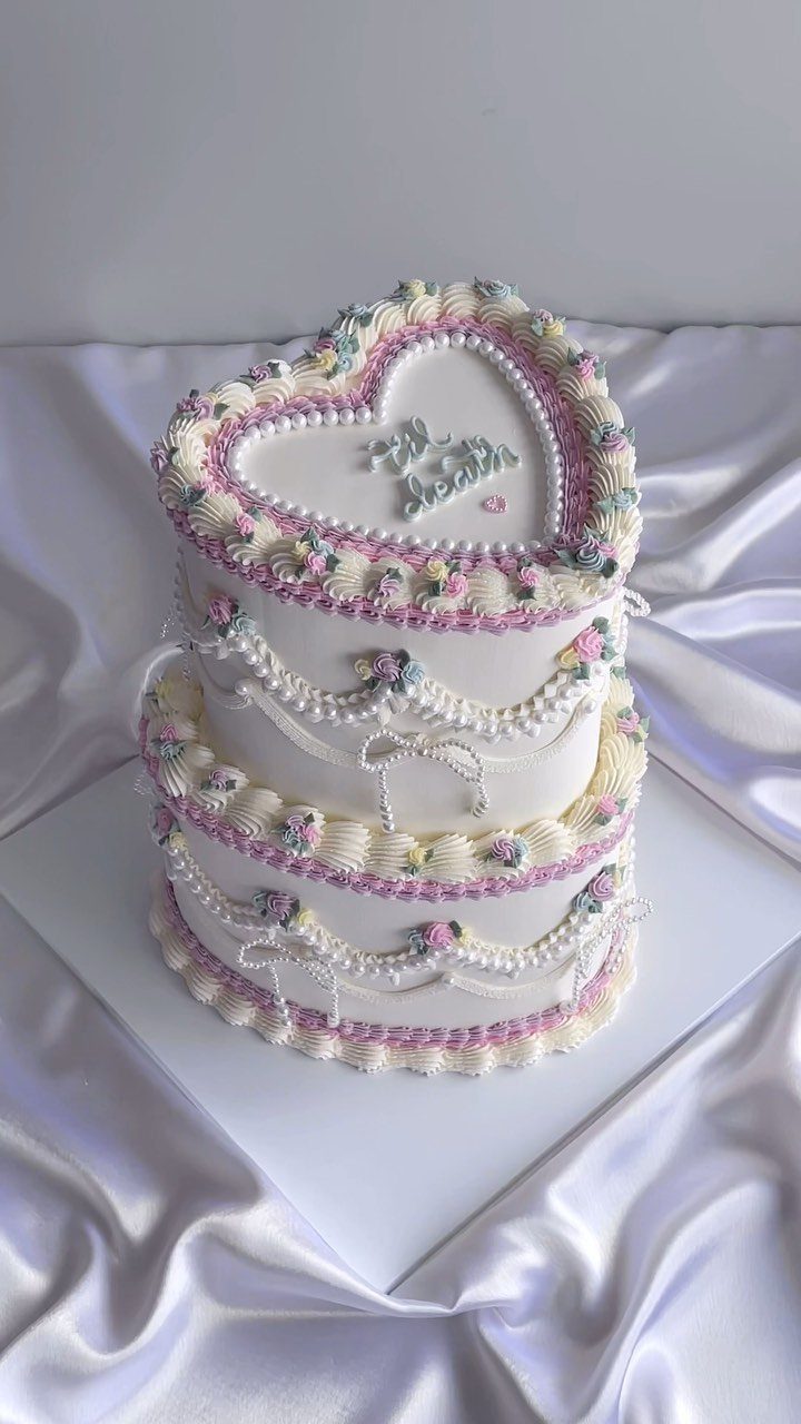 Heart-shaped two-tier white cake with pastel buttercream piping on white satin fabric.