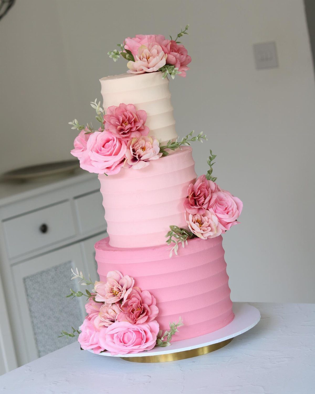 Three-tier pink ombre buttercream cake decorated with fresh pink roses and green leaves.