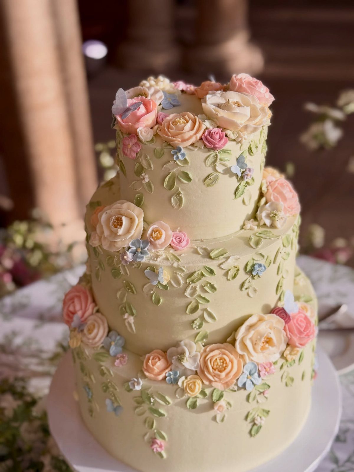 Three-tiered pale yellow cake decorated with buttercream flowers and green leaf vines.