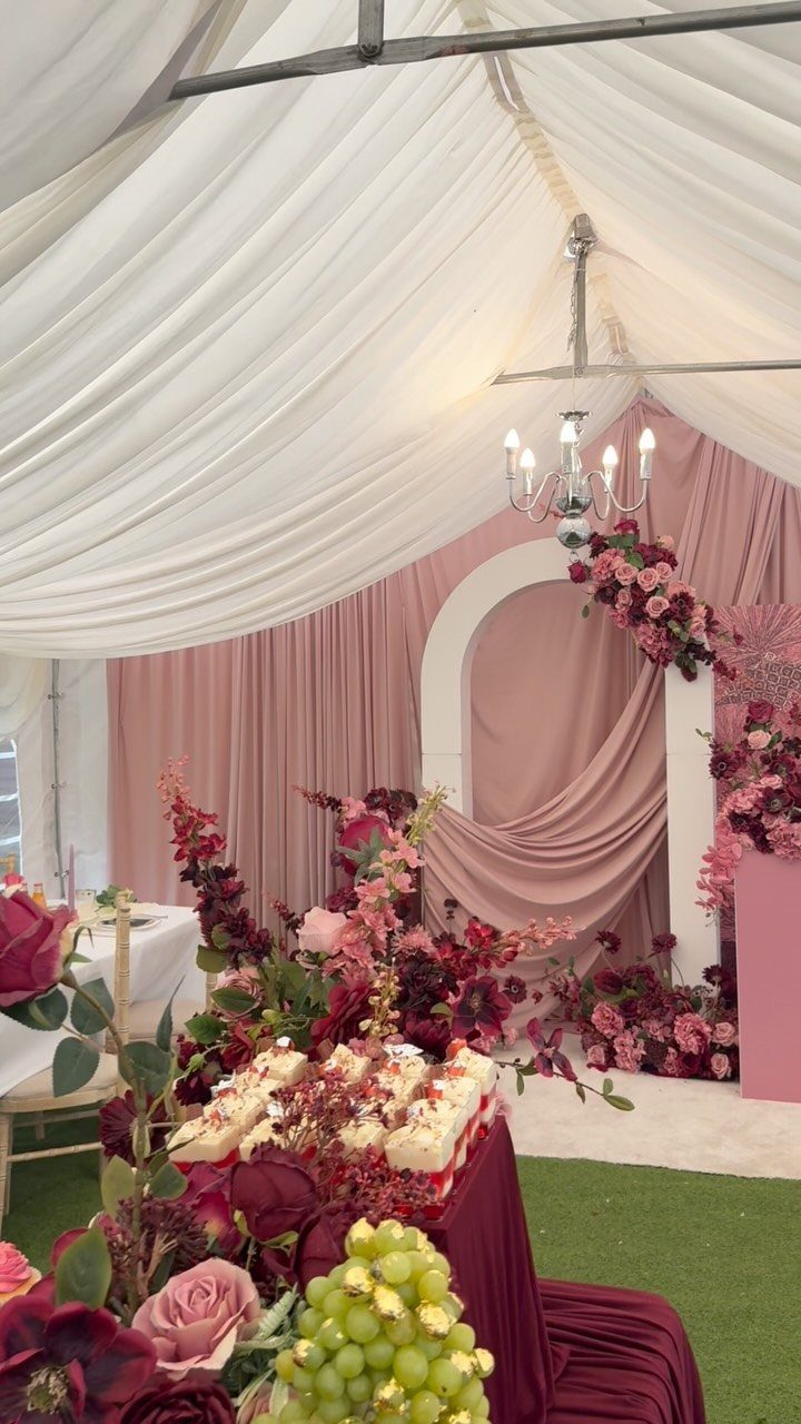 Blush pink and burgundy wedding decor with floral arrangements in a draped tent.
