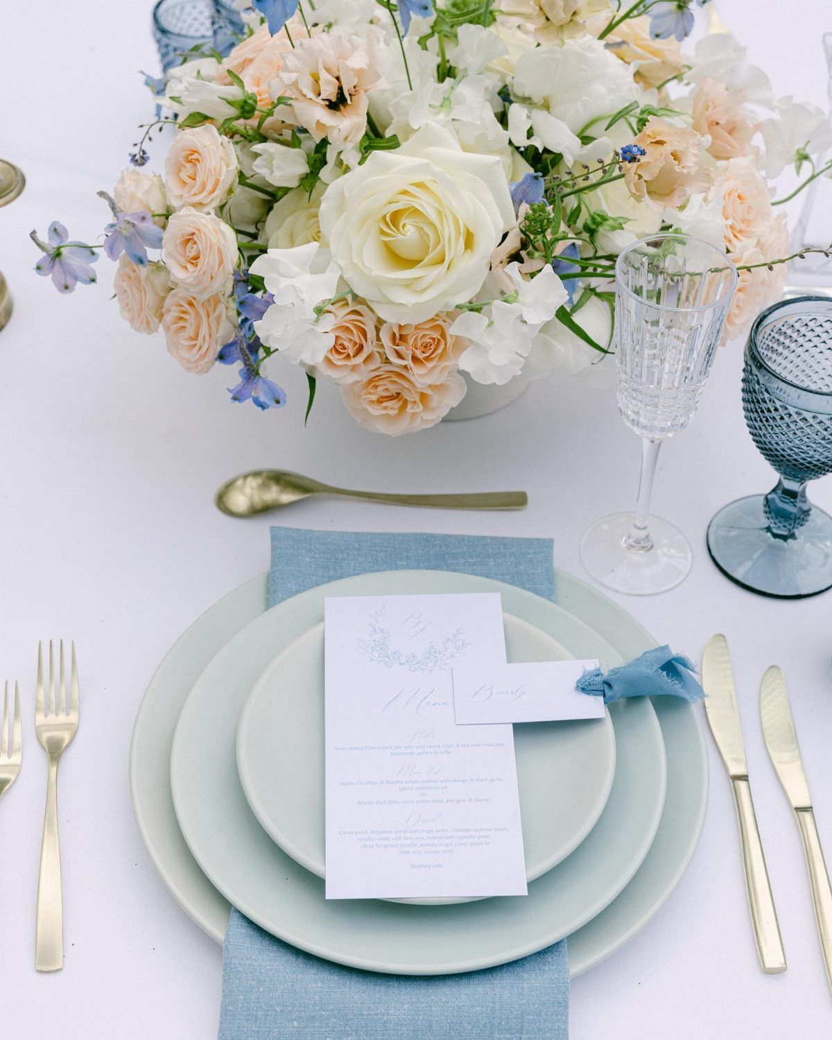 Wedding table setting with peach and white flowers, blue plates, and gold cutlery.
