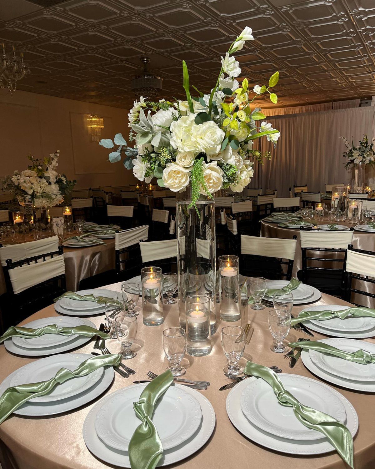 Champagne tablecloth with sage green napkins and white floral centerpiece at wedding reception