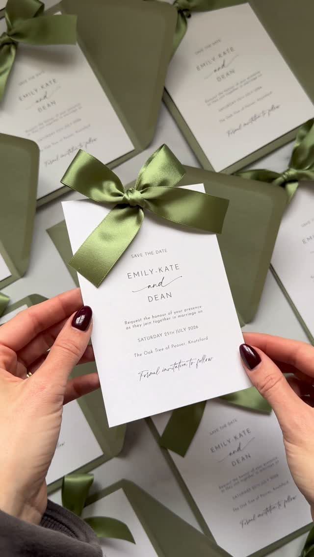 White wedding invitation with a green satin bow held by a person's hands.