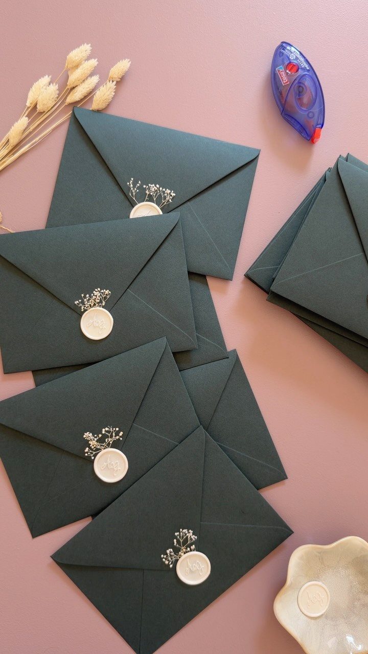 Dark green envelopes with white wax seals and dried flowers on pink background