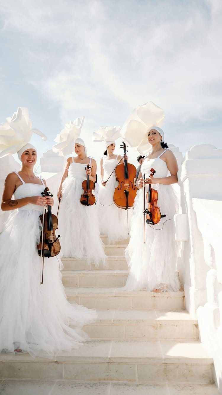 Four women in white gowns posing with string instruments on white stone stairs.