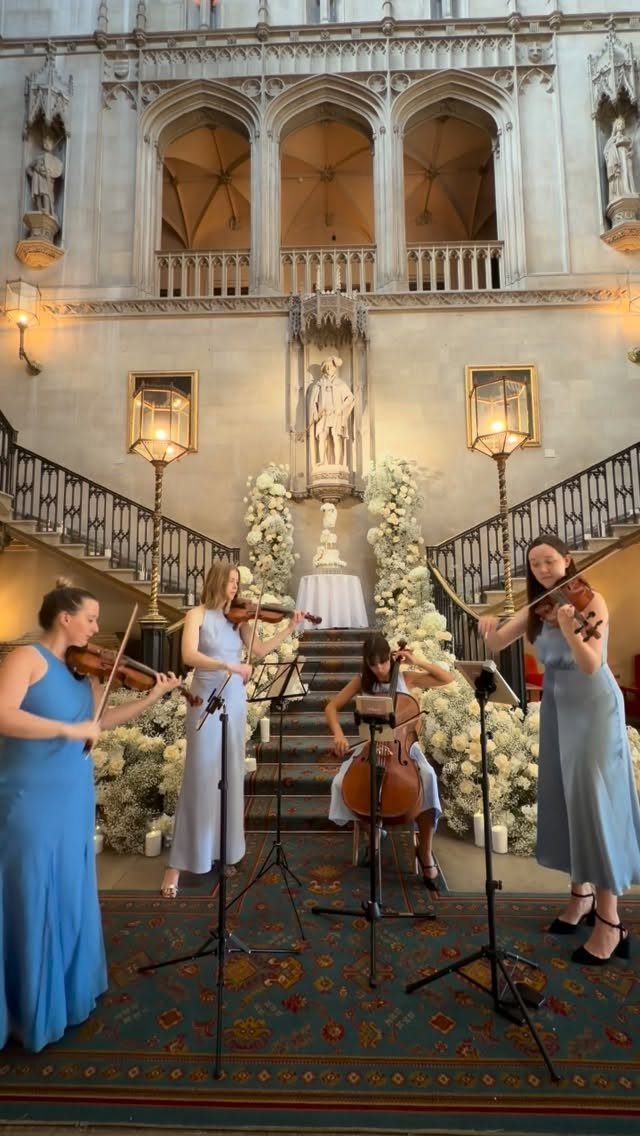 String quartet in blue dresses performing in a grand hall with white flowers.