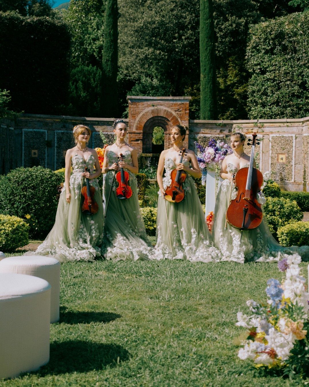 Four women in green gowns playing string instruments in a lush garden setting.
