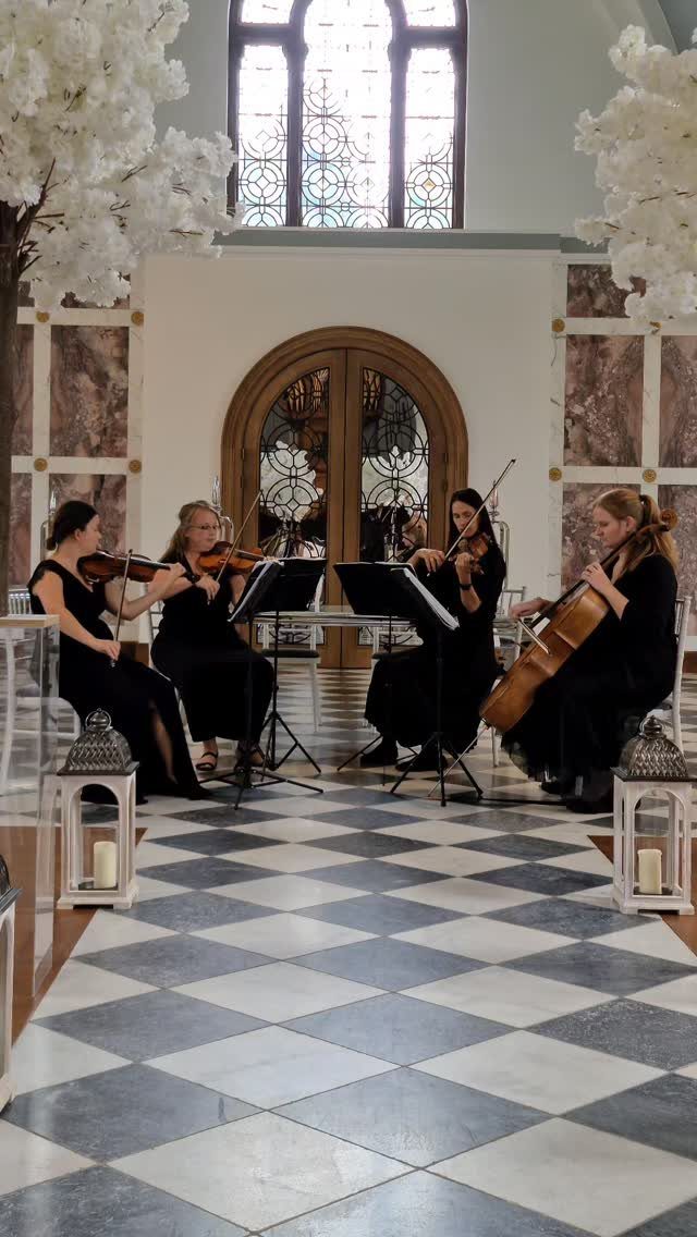 String quartet in black dresses playing instruments in a bright, elegant wedding venue.