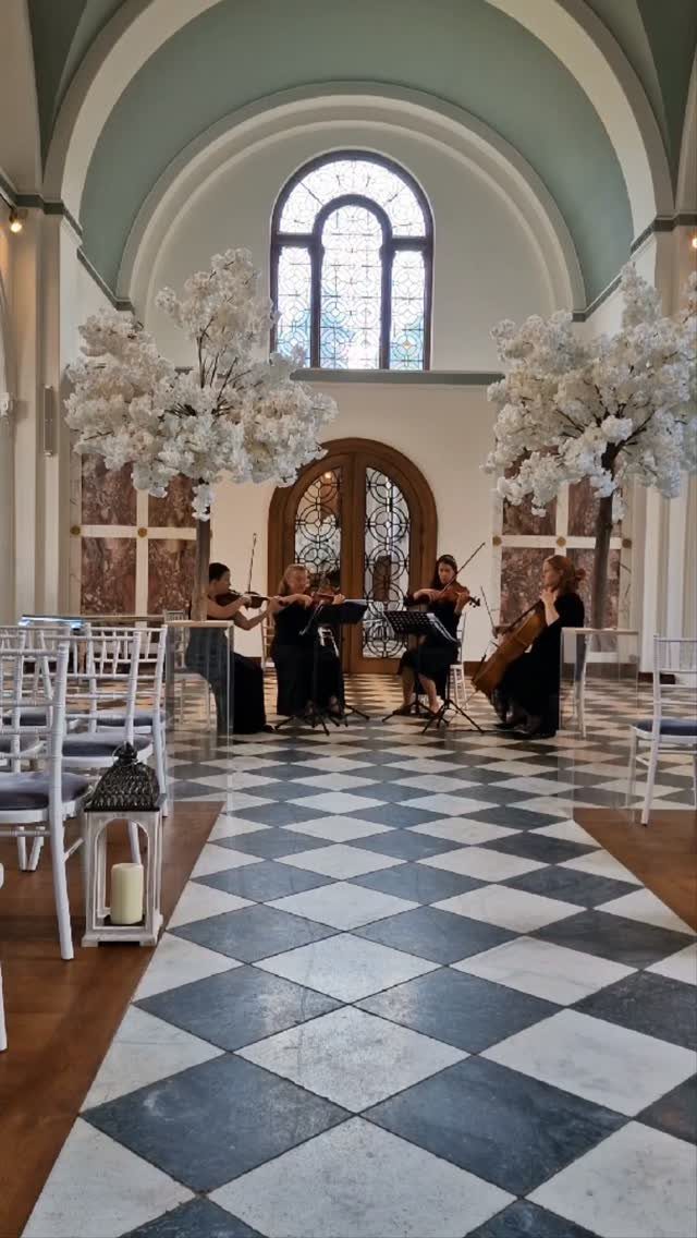String quartet playing instruments in a bright hall with checkered floors and white trees.