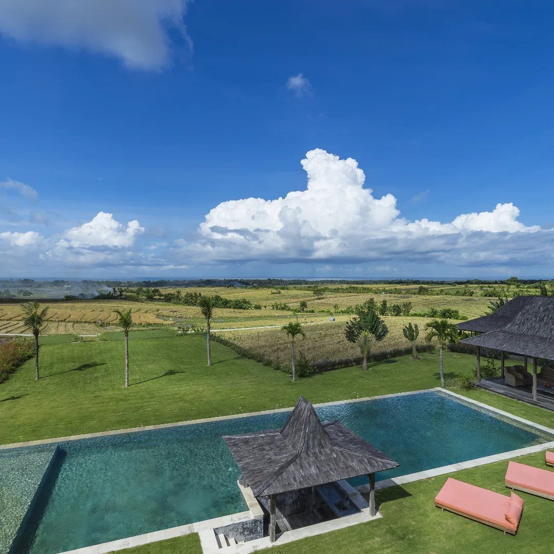 Luxury resort pool and gazebo overlooking expansive green Balinese rice fields under blue sky.