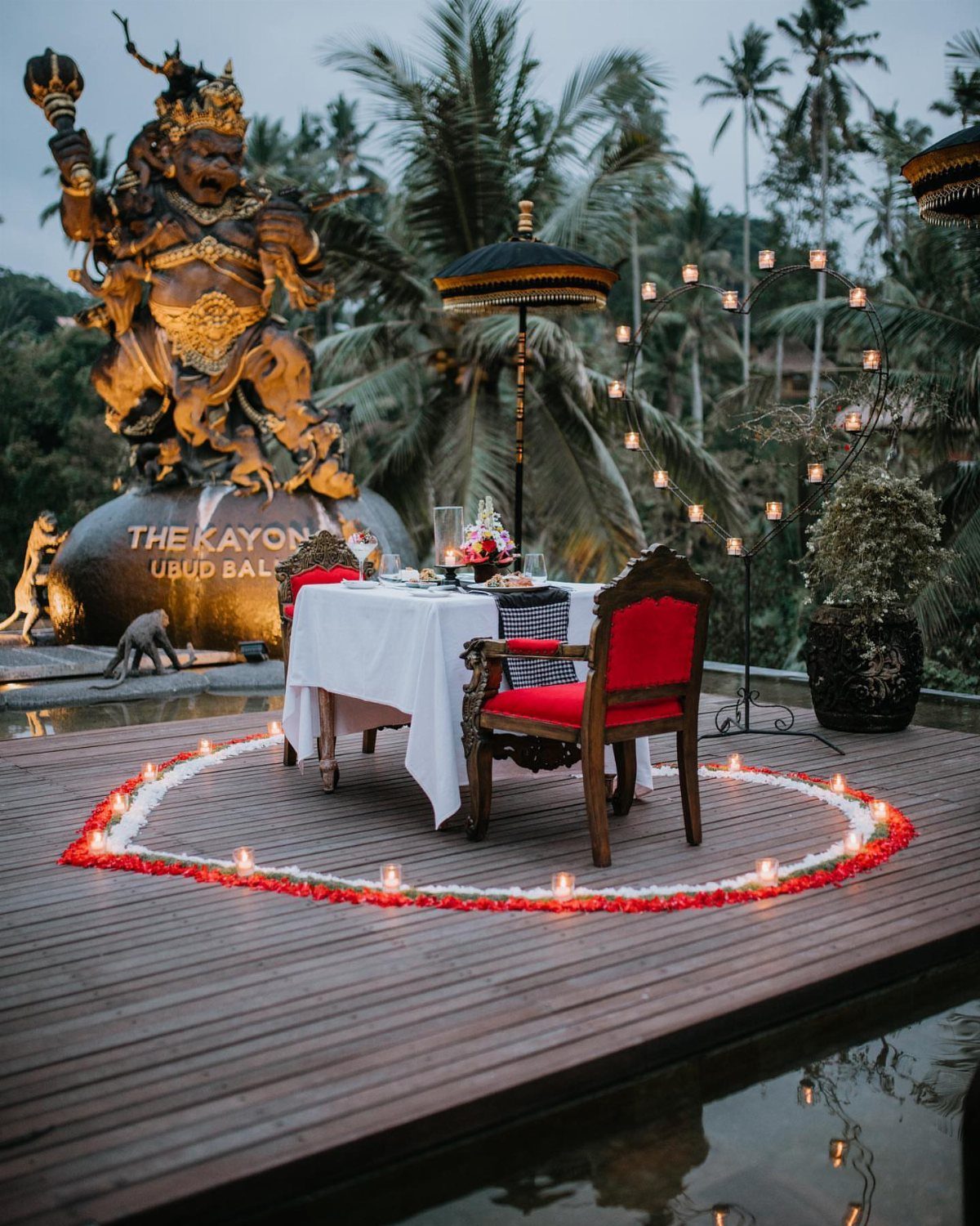Romantic dinner table with red chairs and flower petals near a golden statue.