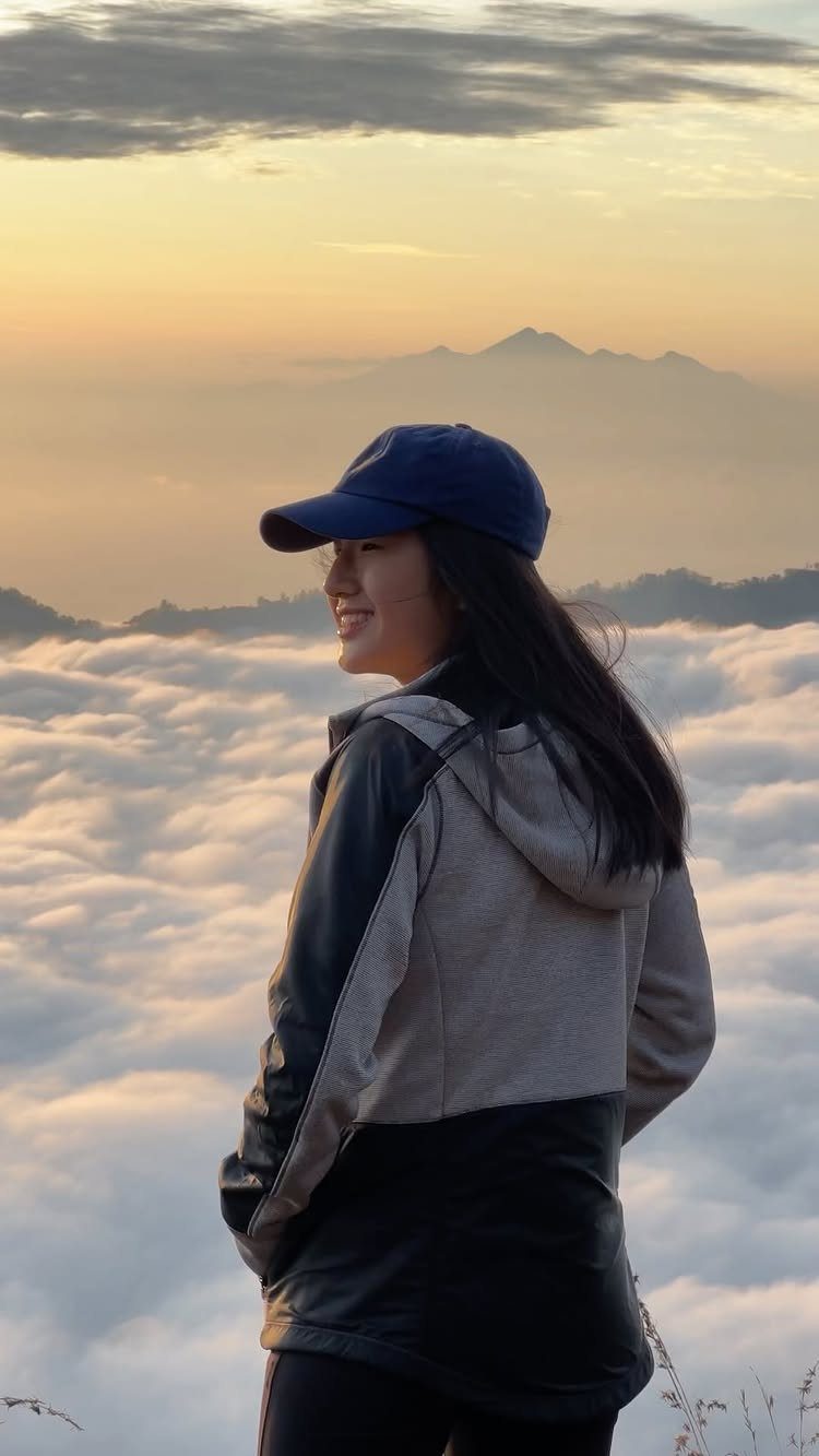 Woman in a cap overlooking a sea of clouds and distant mountains at sunrise.