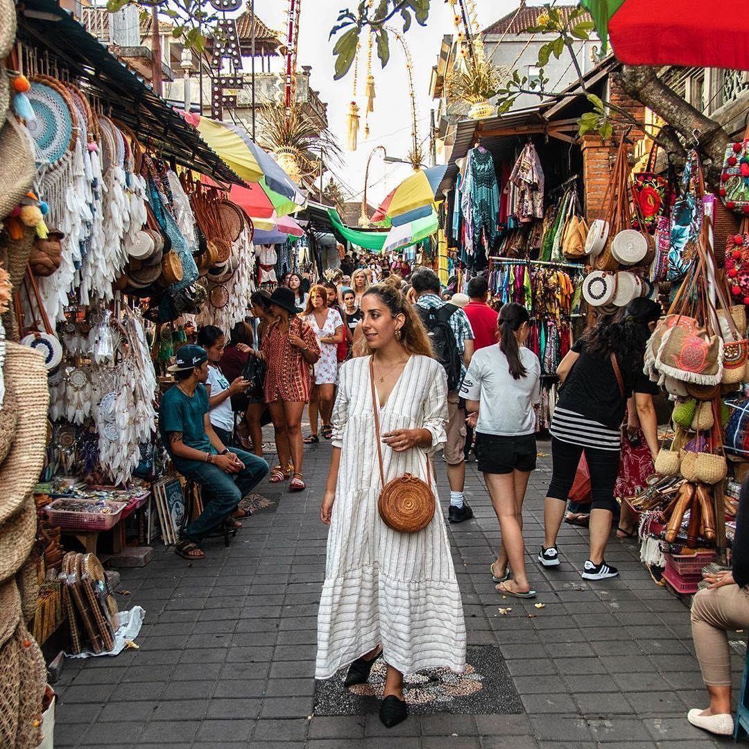 Woman in white dress walking through a vibrant Ubud market with woven bags.