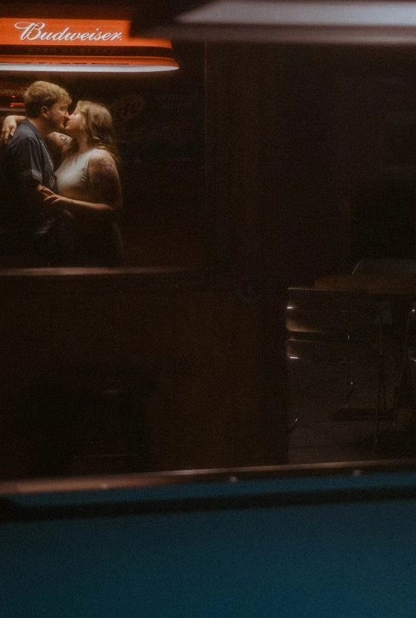 Couple embracing in a dark bar under a glowing red Budweiser sign.