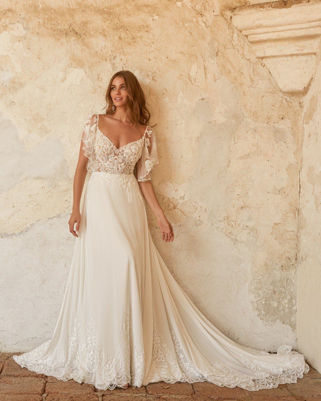 Woman in an ivory lace wedding dress posing against a textured stone wall.