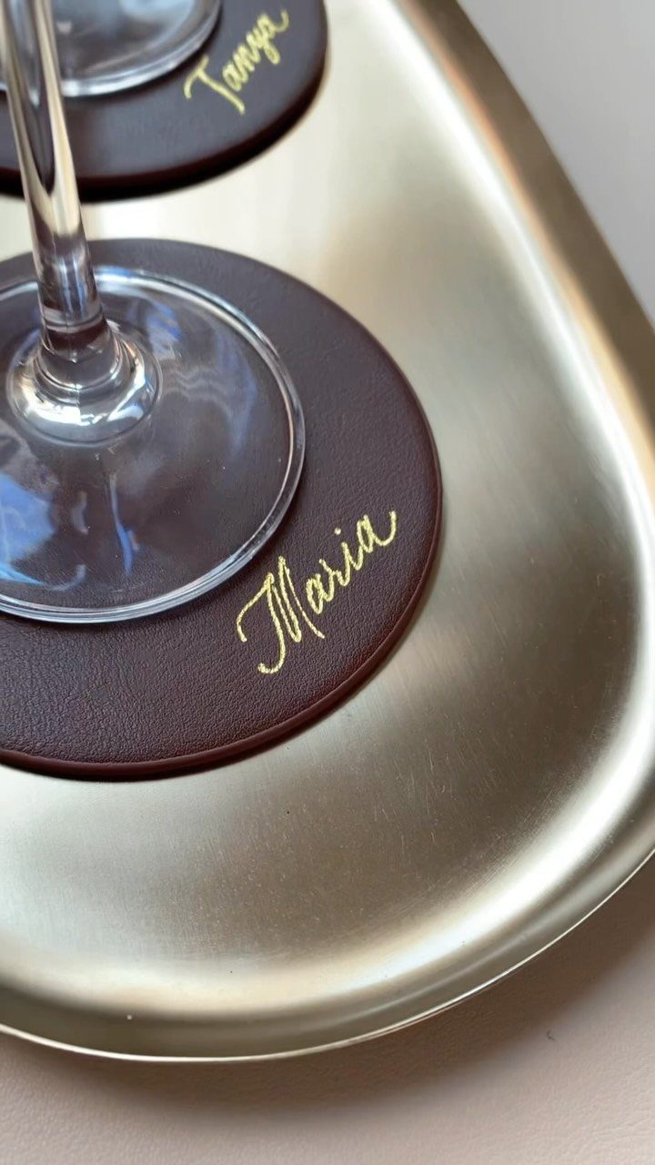 Dark brown leather coasters with gold cursive names on a gold metallic tray.