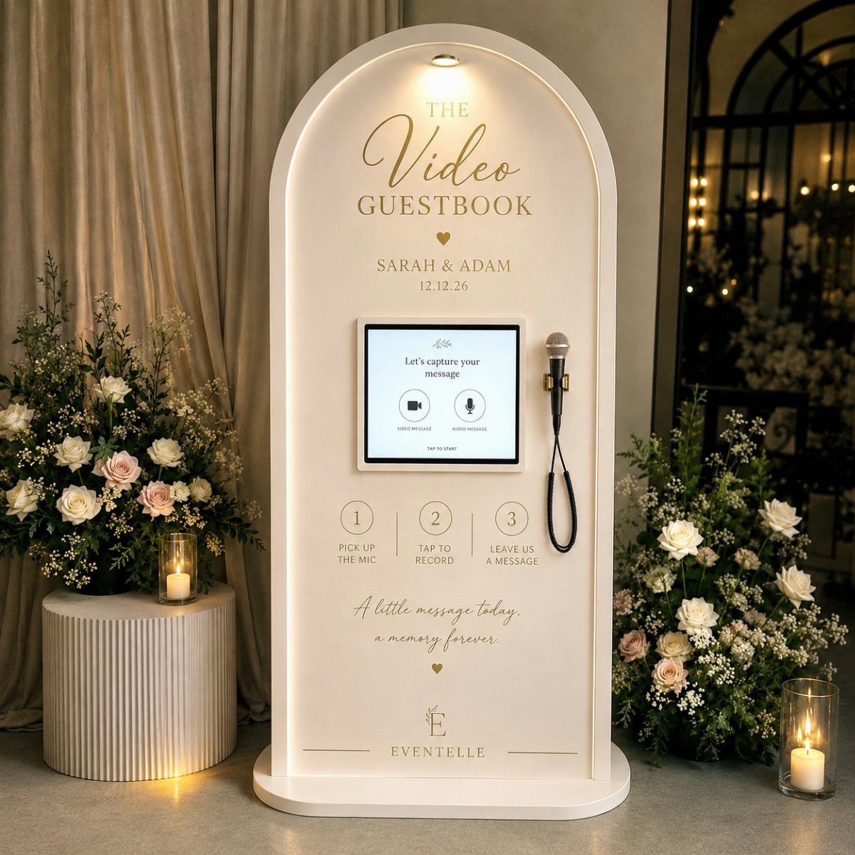 White arched video guestbook kiosk with a screen and microphone surrounded by flowers.