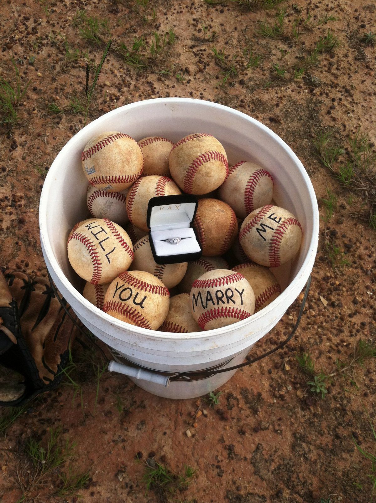 White bucket of baseballs with a marriage proposal message and an engagement ring.