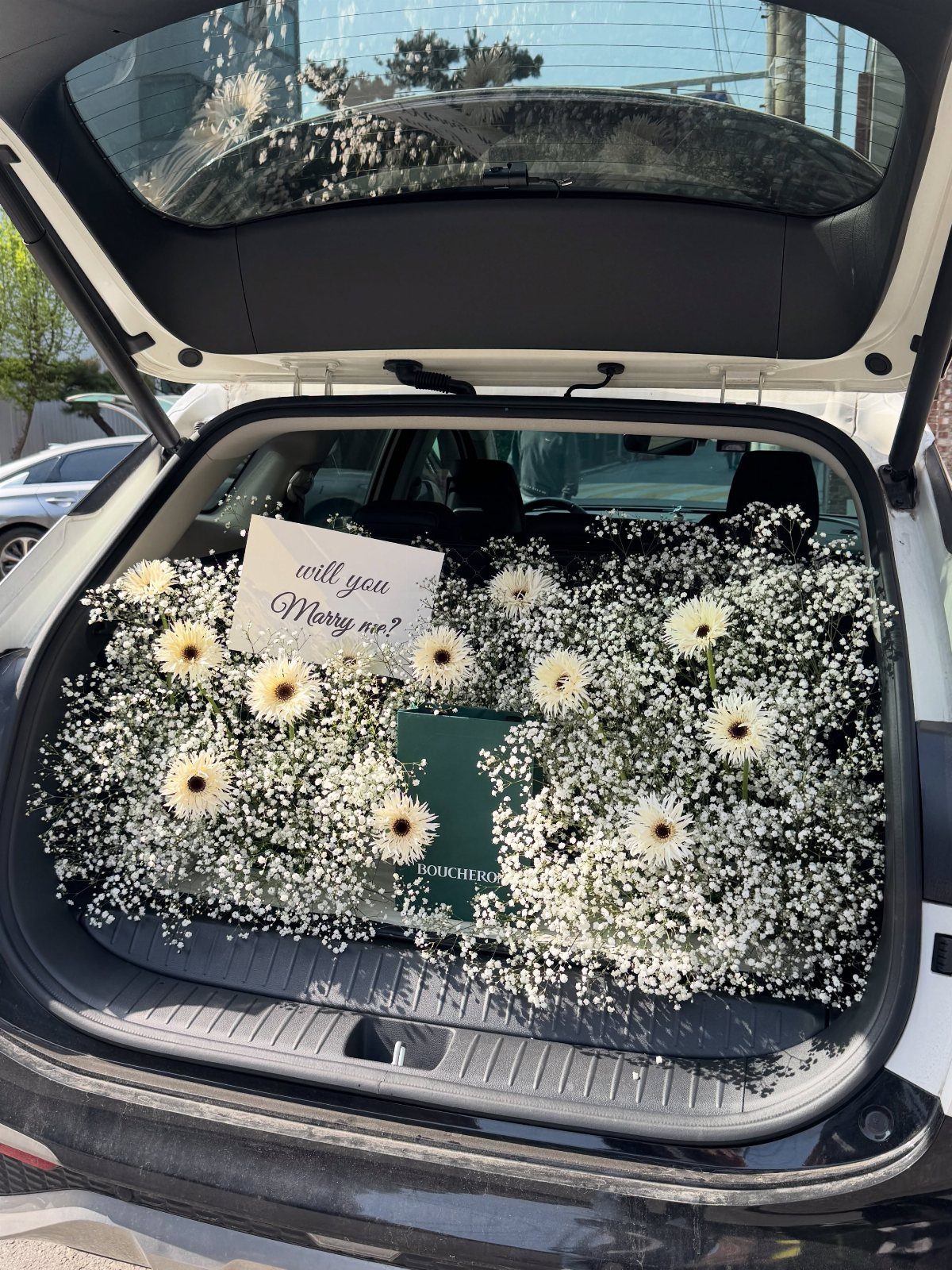 Car trunk filled with white flowers and a will you marry me card