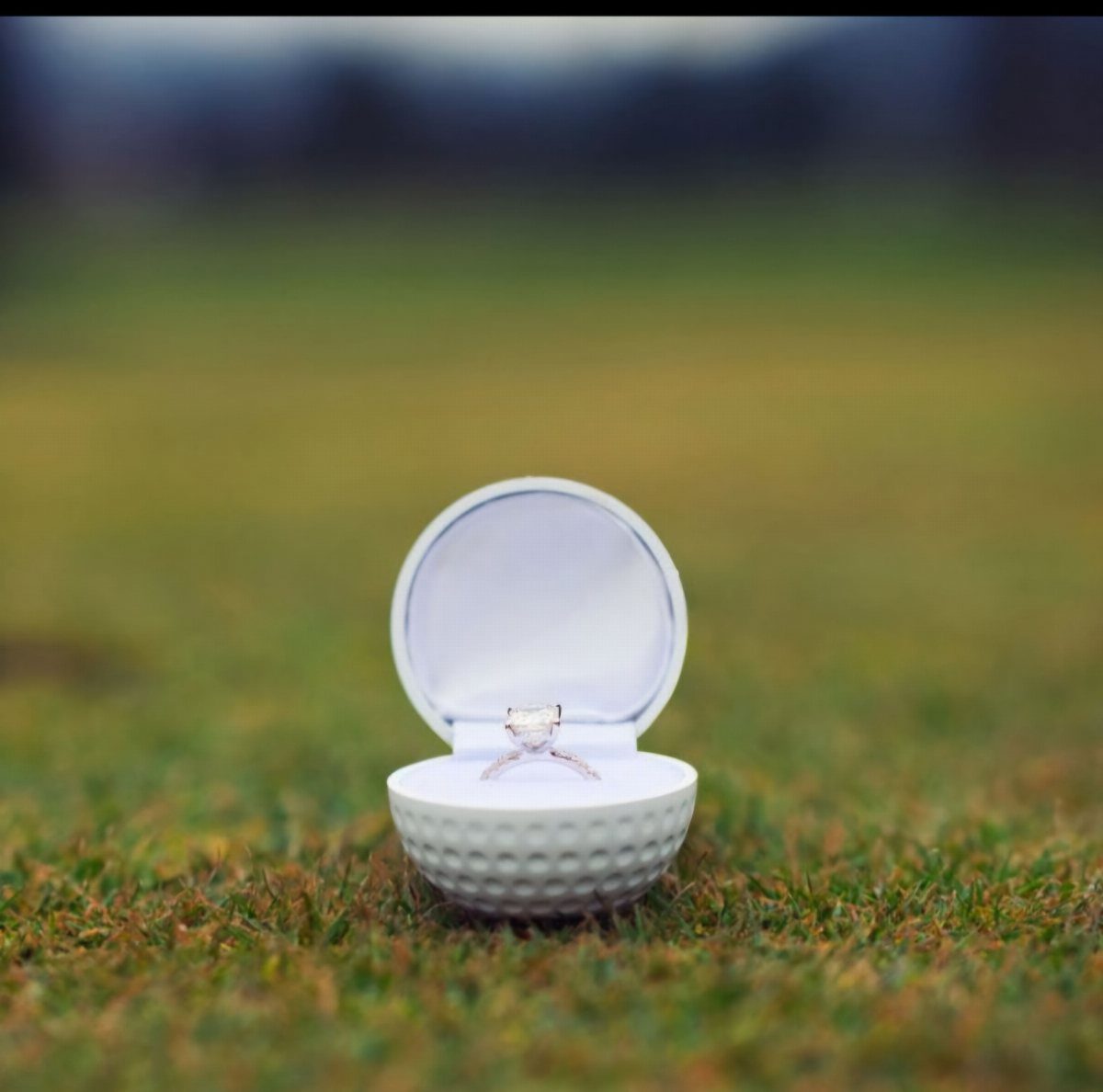 Diamond engagement ring inside a golf ball ring box on green grass.