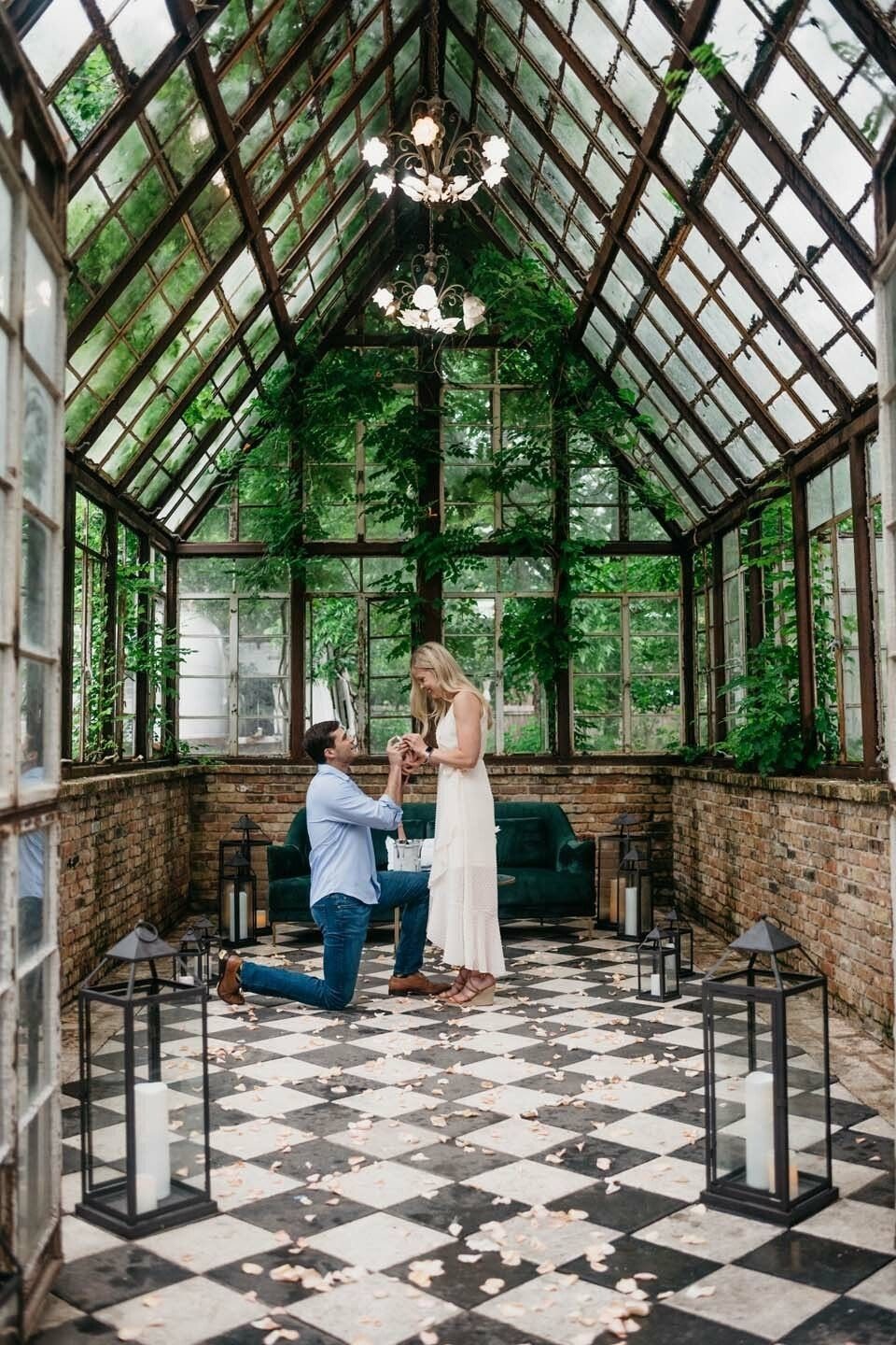 Man kneeling to propose to a woman inside a glass greenhouse with lanterns.
