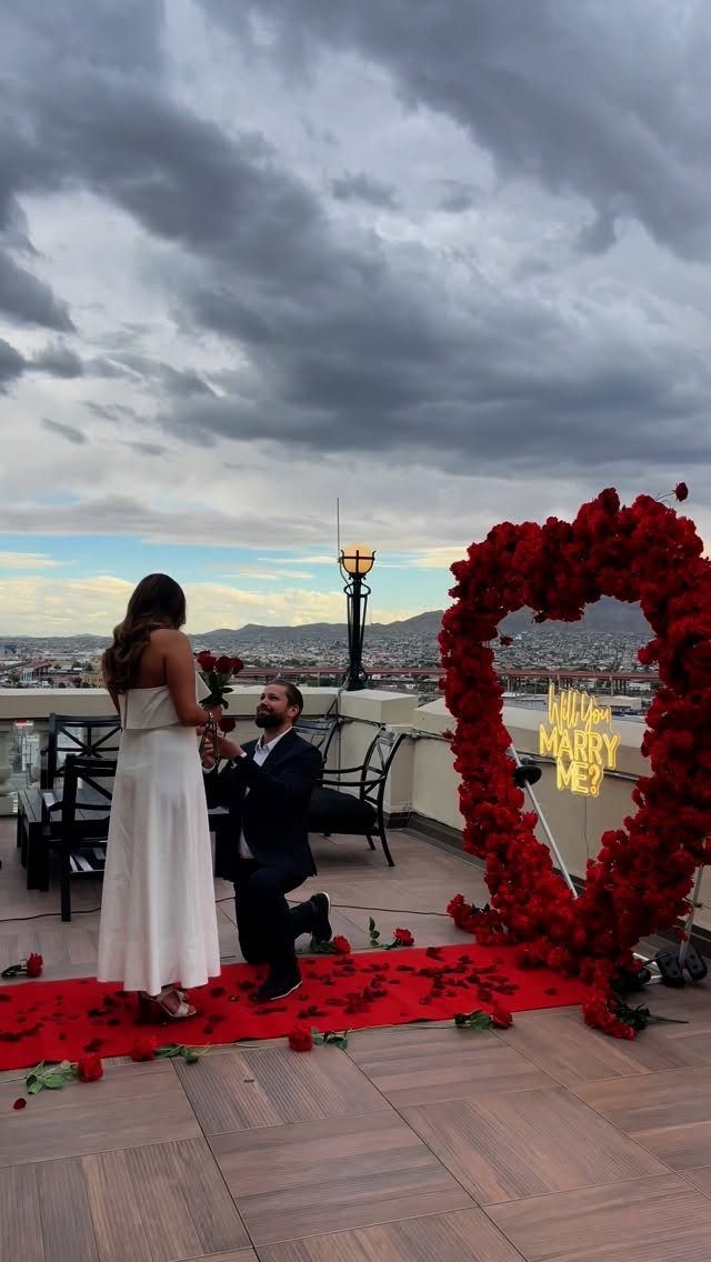 Man kneeling on a rooftop proposing to a woman beside a rose heart arch.